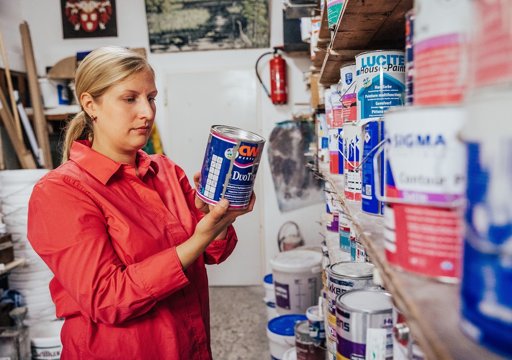 Eine Frau in einem roten Hemd steht in einem Lagerraum und untersucht eine Farbdose in einem Regal, das mit verschiedenen Farbdosen und Eimern gefüllt ist. Die Regale sind geordnet und der Raum scheint eine Werkstatt oder ein Vorratsraum zu sein.