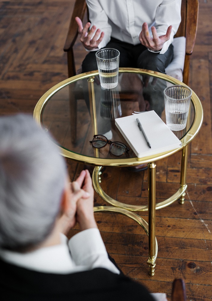 Zwei Menschen sitzen sich an einem runden Glastisch gegenüber, auf dem zwei Gläser Wasser, ein Notizbuch, ein Stift und eine Brille stehen. Während der eine beim Reden gestikuliert, hört Sabine Logemann aufmerksam zu und schafft so einen Raum für offene Selbstentfaltung.