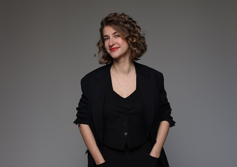 Eine Frau mit lockigem, schulterlangem Haar, die einen schwarzen Blazer und ein schwarzes Oberteil trägt, steht vor einem schlichten grauen Hintergrund und lächelt, die Hände in den Taschen.