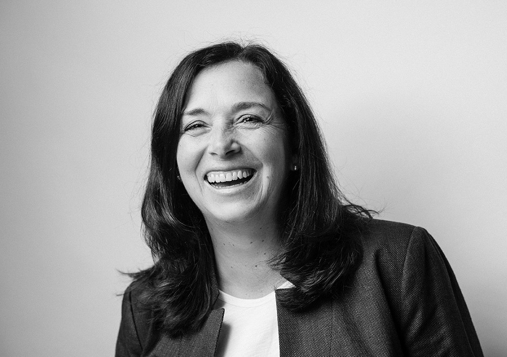 Eine Frau mit langen dunklen Haaren, die einen Blazer und ein helles Oberteil trägt, lächelt breit, während sie vor einem schlichten Hintergrund etwas aus dem Bild herausschaut. Das Bild ist in schwarz-weiß gehalten.