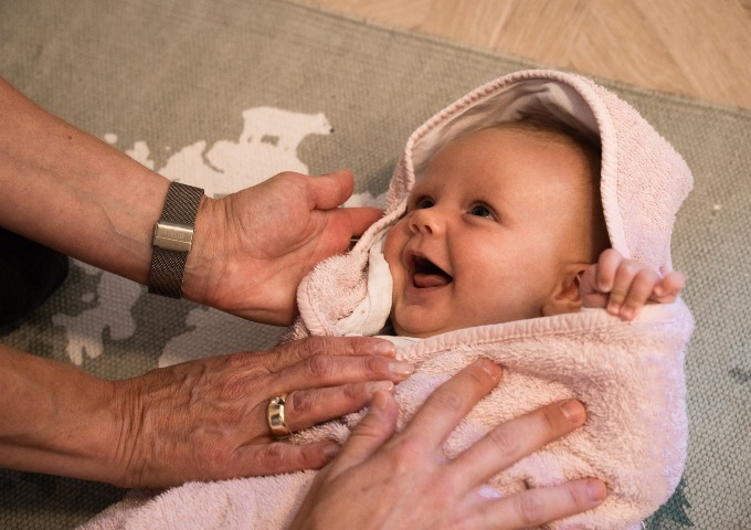 Ein lächelndes, in ein rosafarbenes Handtuch eingewickeltes Baby blickt auf, während ein Erwachsener sanft sein Gesicht und seine Brust berührt und so eine warme und liebevolle Atmosphäre schafft.