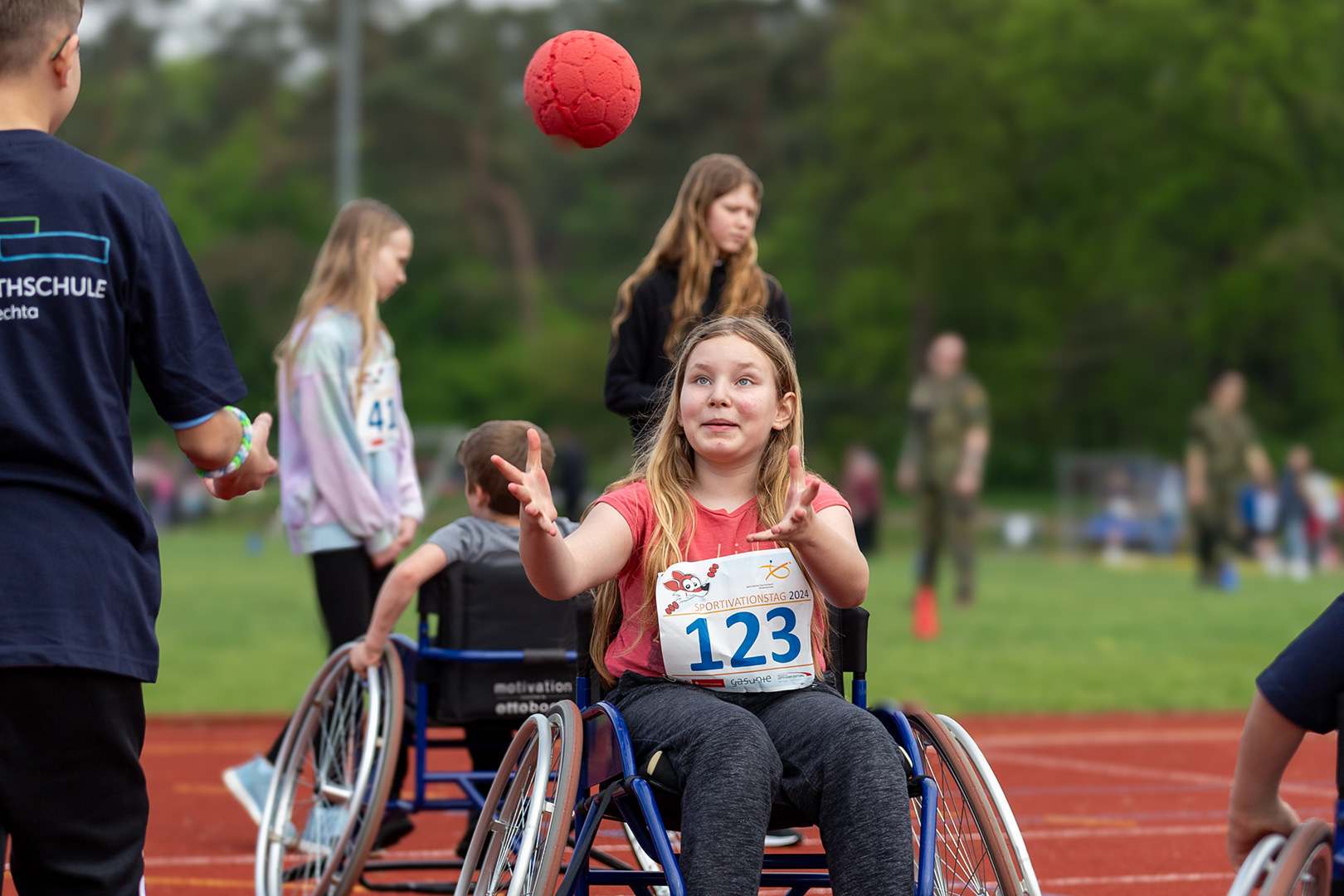 Ein Mädchen im Rollstuhl, das die Startnummer 123 trägt, fängt einen roten Ball auf einer Sportbahn. Andere Kinder und Erwachsene, einige auch im Rollstuhl, nehmen an Aktivitäten teil.