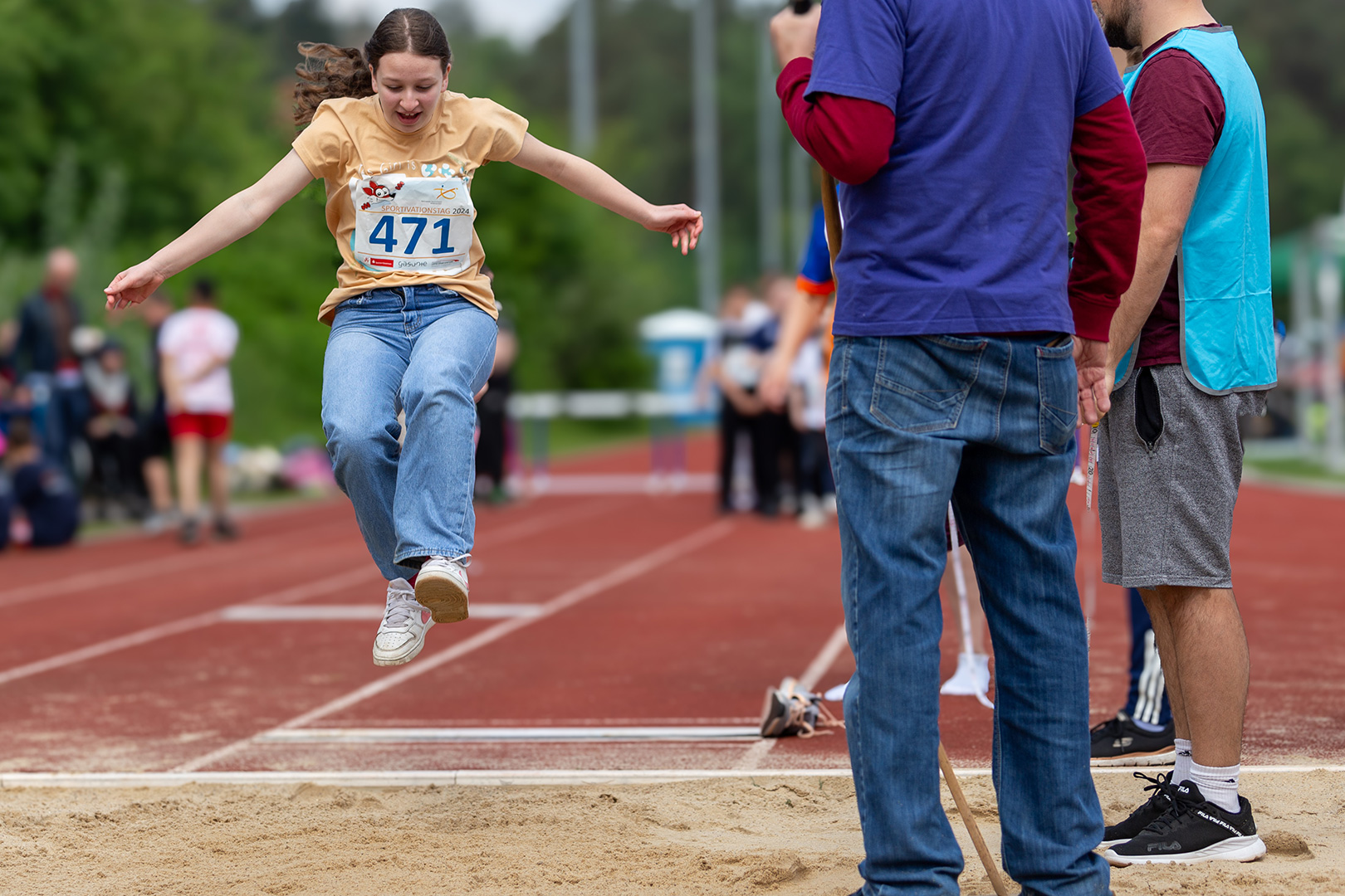 Eine junge Sportlerin in gelbem Hemd und Jeans, mitten in der Luft beim Weitsprung auf einer Bahn, mit Sand unter ihr und Offiziellen, die in der Nähe zuschauen. Zuschauer und Grünflächen füllen den Hintergrund.