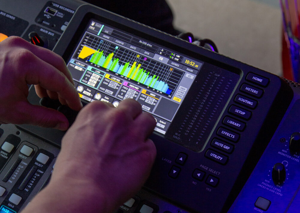 Nahaufnahme von Händen, die Bedienelemente an einem Audiomischpult mit einem Touchscreen einstellen, auf dem Audiopegel, Equalizer-Einstellungen und verschiedene farbige Klangwellenformen angezeigt werden.