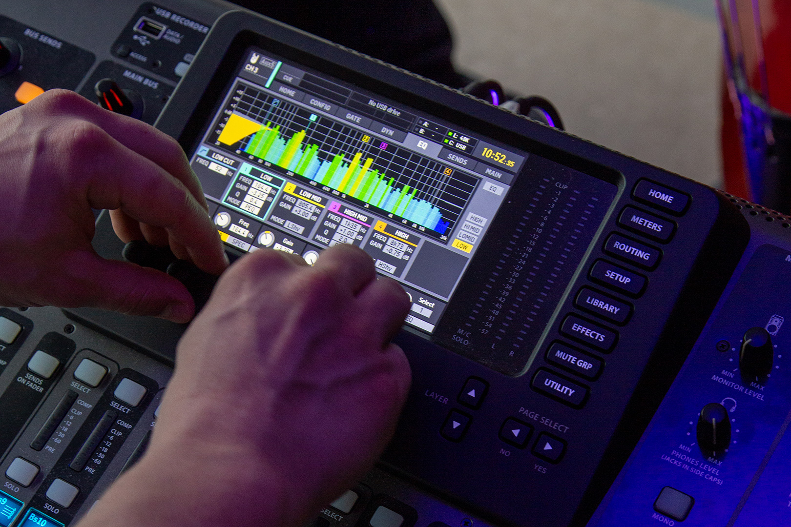 Nahaufnahme von Händen, die Bedienelemente an einem Audiomischpult mit einem Touchscreen einstellen, auf dem Audiopegel, Equalizer-Einstellungen und verschiedene farbige Klangwellenformen angezeigt werden.