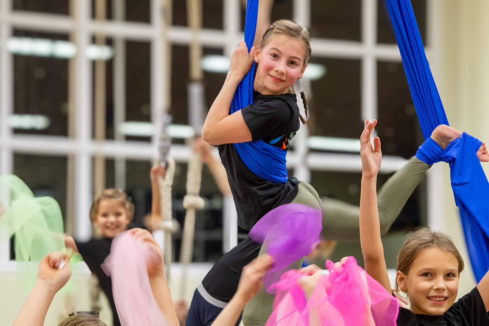 Junge Mädchen führen in einer Halle Luftakrobatik auf; ein Mädchen hängt an einem blauen Stoff und lächelt in die Kamera, während andere jubeln und bunte Tücher hochhalten. Helle, lebendige und energiegeladene Atmosphäre.