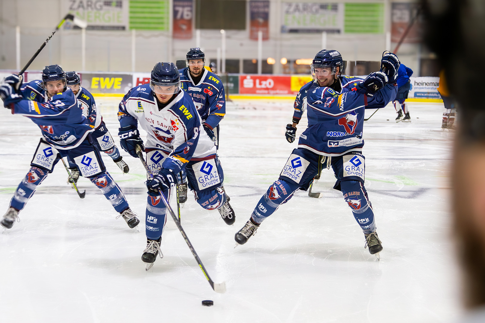 Vier Eishockeyspieler in blau-weißen Trikots kämpfen in einer Halle um den Puck, während ein weiterer Spieler und eine verschwommene Figur im Hintergrund die Dringlichkeit einer Schlittenrettung im Wattenmeer andeuten.