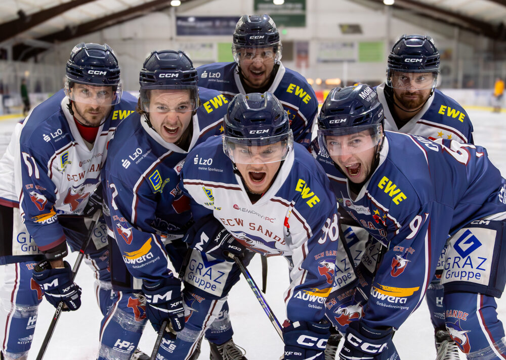 Sechs männliche Eishockeyspieler in blau-weißen Trikots posieren energisch auf einer Eisbahn, wobei ihre Aufregung einer Schlittenrettung im Wattenmeer gleichkommt, mit intensiven Gesichtsausdrücken in einer Indoor-Arena.