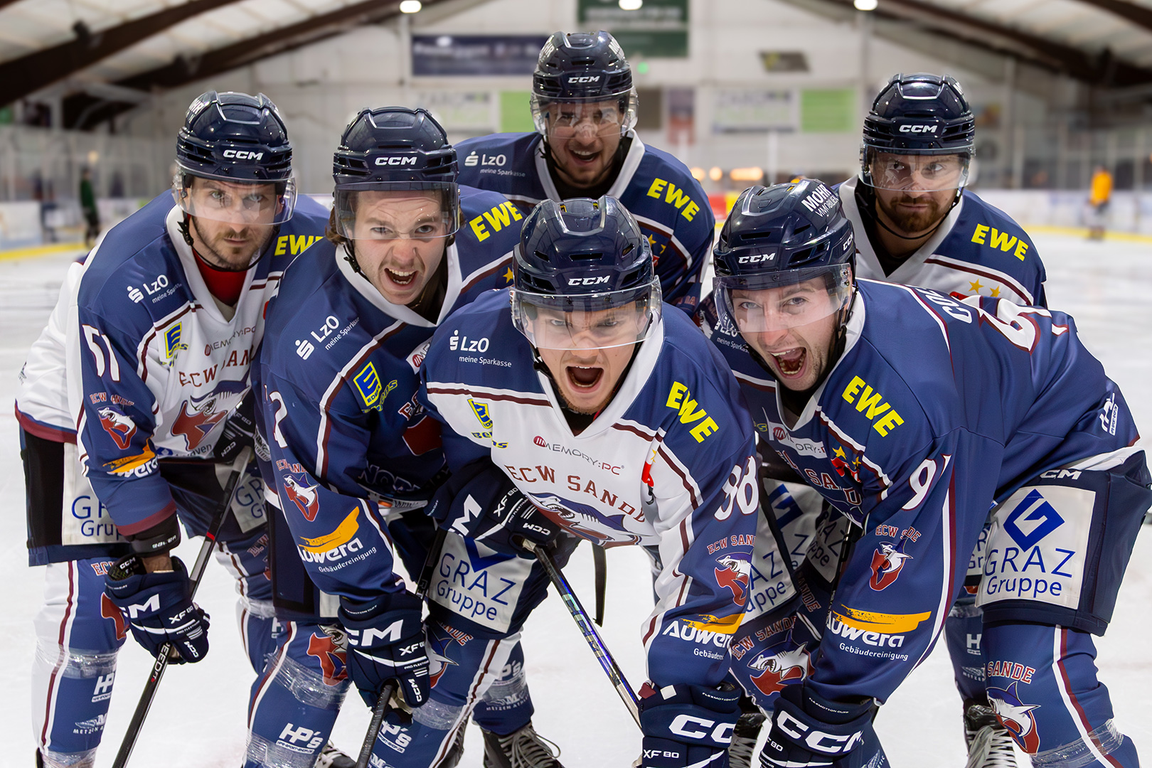 Sechs männliche Eishockeyspieler in blau-weißen Trikots posieren energisch auf einer Eisbahn, wobei ihre Aufregung einer Schlittenrettung im Wattenmeer gleichkommt, mit intensiven Gesichtsausdrücken in einer Indoor-Arena.