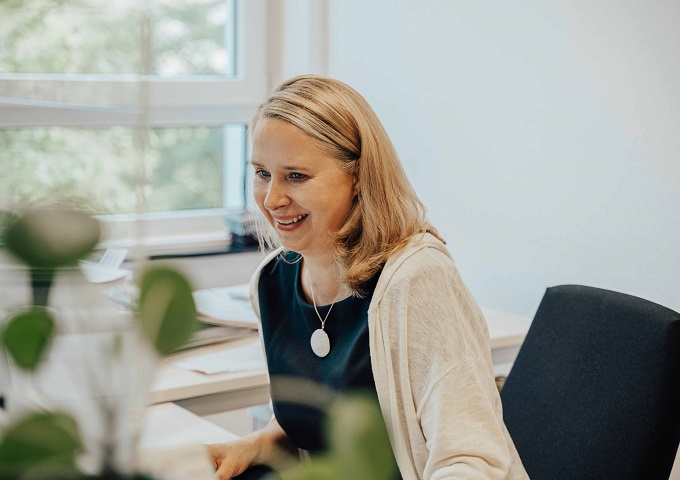 Eine Frau mit blondem Haar, schwarzem Oberteil und weißer Strickjacke, sitzt lächelnd an einem Schreibtisch in einem hellen Büro mit Fenster und Pflanzen in der Nähe.