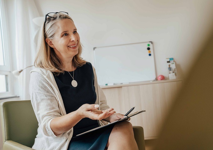 Eine lächelnde Frau sitzt in einem Bürostuhl, hält ein Klemmbrett in der Hand und gestikuliert mit ihrer Hand. Sie hat schulterlanges blondes Haar, trägt eine Brille auf dem Kopf, und im Hintergrund ist ein Whiteboard zu sehen.