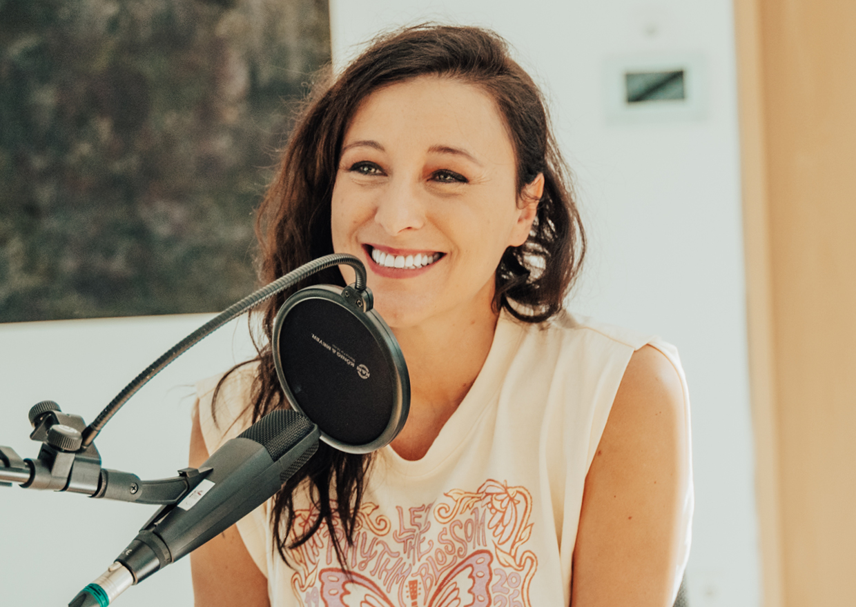 Annie Heger, eine lächelnde Frau mit langen braunen Haaren, sitzt in einem ärmellosen Hemd mit Schmetterlingsmuster vor einem Mikrofon und ist bereit, in einem hellen Innenraum Aufnahmen zu machen oder zu sprechen.