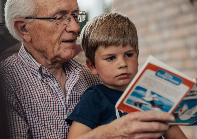 Ein älterer Mann mit Brille sitzt neben einem kleinen Jungen, beide lesen ein Buch mit Vögeln auf dem Einband. Der Mann trägt ein kariertes Hemd und der Junge ein blaues T-Shirt. Sie wirken konzentriert und beschäftigt.