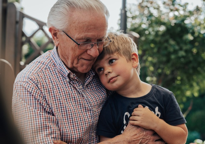 Ein älterer Mann mit Brille und weißem Haar umarmt liebevoll einen kleinen Jungen, der seinen Kopf auf die Schulter des Mannes stützt. Sie sind im Freien, umgeben von Grün, und beide scheinen zufrieden und einander nahe zu sein.