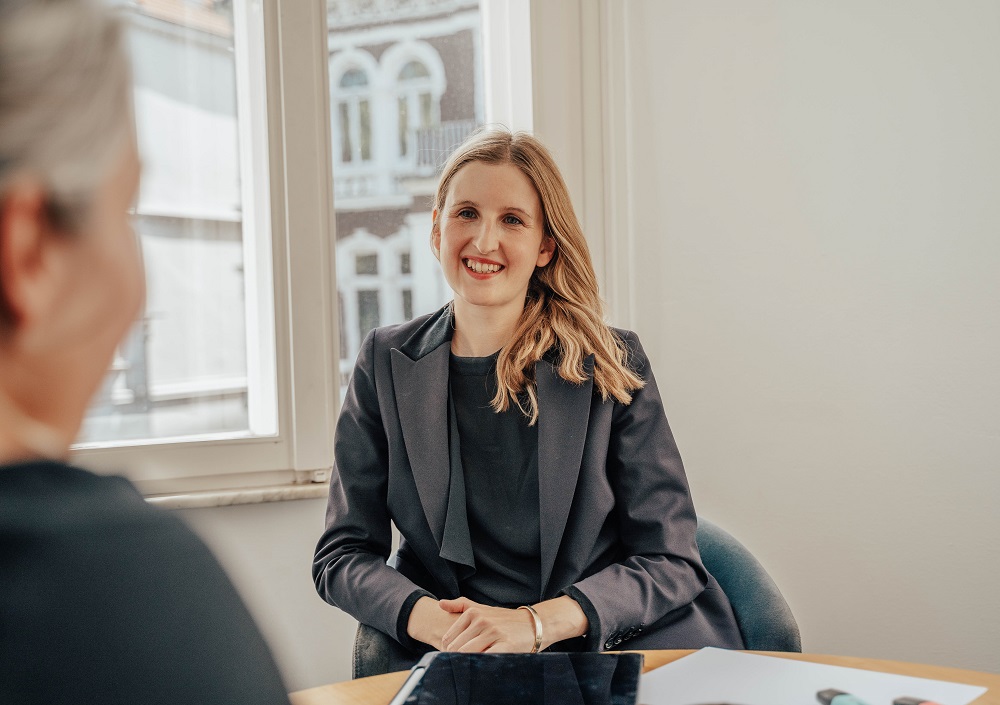 Eine Frau mit langen blonden Haaren, die einen dunklen Blazer trägt und lächelt, sitzt an einem Tisch in einem hellen Büro und blickt auf eine andere Person, die mit dem Rücken zur Kamera steht.