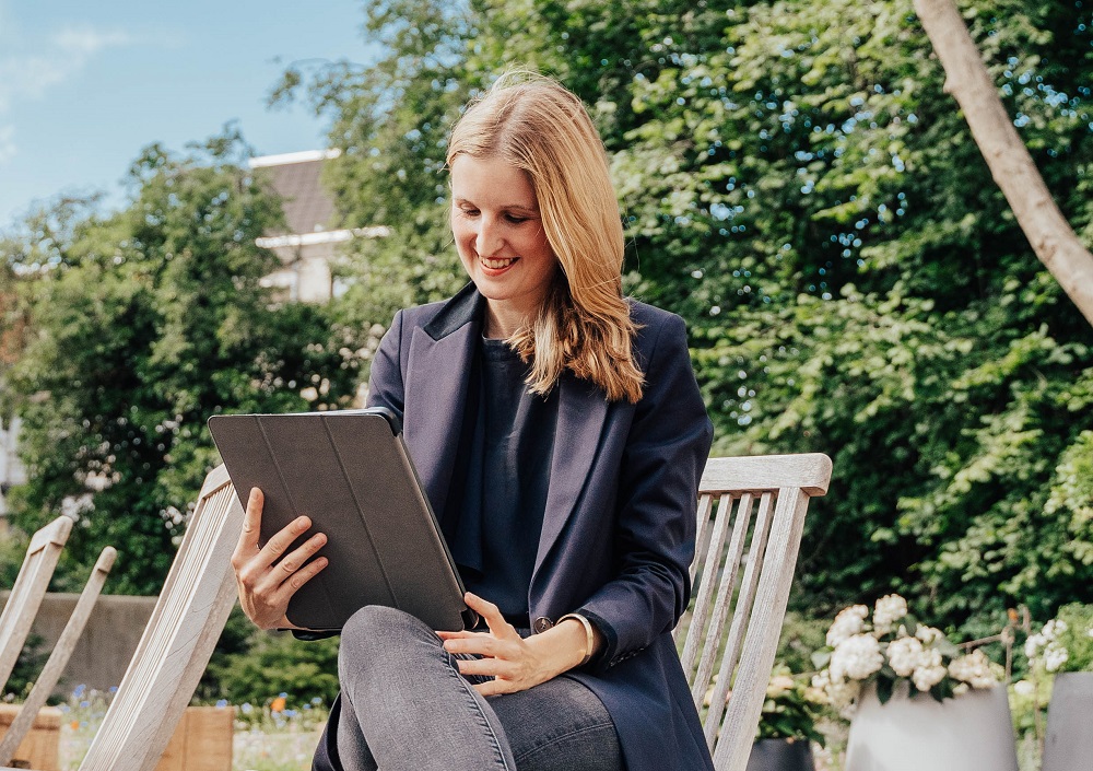 Eine Frau sitzt im Freien auf einem Holzstuhl und lächelt, während sie ein Tablet benutzt. Sie trägt einen dunklen Blazer und Jeans, im Hintergrund sind Grünpflanzen und Blumen zu sehen.