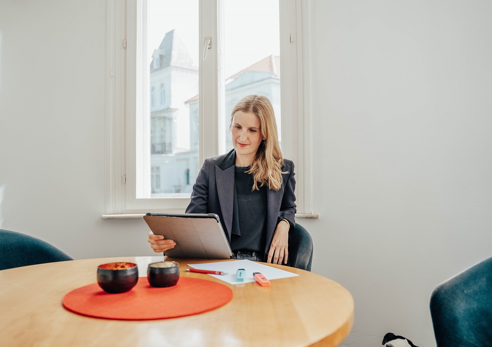 Eine Frau sitzt an einem runden Tisch mit Papieren, Stiften und Schalen vor sich und schaut auf eine Tafel. Sie befindet sich in einem hellen Raum mit zwei großen Fenstern hinter ihr.