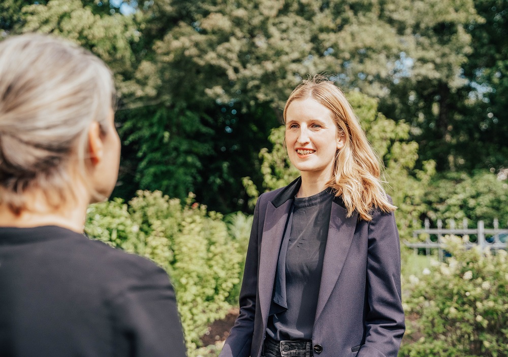 Zwei Frauen stehen im Freien und führen ein Gespräch. Eine Frau mit blondem Haar schaut in die Kamera und lächelt, während die andere von hinten zu sehen ist. Im Hintergrund sind üppige Grünflächen und Bäume zu sehen.