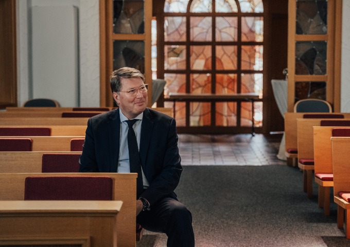 Ein Mann im Anzug und mit Brille sitzt allein auf einer Holzbank in einer Kirche, lächelt und schaut zur Seite. Das Innere der Kirche ist mit rot gepolsterten Sitzen und großen Fenstern ausgestattet, durch die natürliches Licht einfällt.