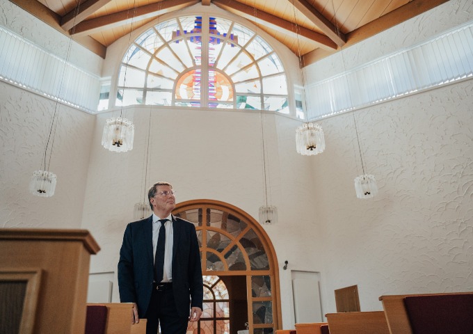 Ein Mann im Anzug steht im Inneren einer Kirche und schaut nach oben. Die Kirche hat weiße Wände, hölzerne Deckenbalken, Hängelampen und ein großes gewölbtes Buntglasfenster über ihm.