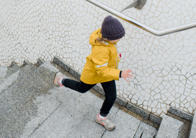 Ein kleines Kind, das einen gelben Regenmantel, eine schwarze Hose und eine lila Strickmütze trägt, rennt eine graue Steintreppe neben einer mosaikgefliesten Wand hinauf.