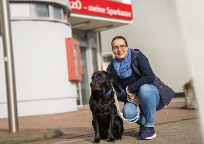 Eine Frau mit Brille und blauem Schal kniet neben einem schwarzen Blindenhund an der Leine vor einem Gebäude mit einem rot-weißen Schild.