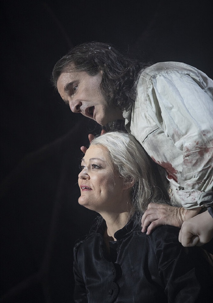 Ein Mann mit langen dunklen Haaren, der ein blutverschmiertes weißes Hemd trägt, beugt sich über eine sitzende Frau mit grauen Haaren, die schwarz gekleidet ist; beide wirken auf einer schwach beleuchteten Bühne sehr emotional.
