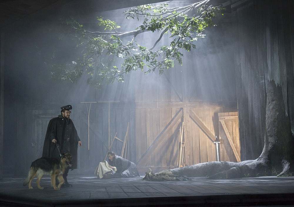 Eine dramatische Bühnenszene zeigt einen dunkel gekleideten Mann mit einem Hund, der neben einer Frau steht, die auf dem Boden unter einem großen Ast kauert, während Lichtstrahlen in eine rustikale, hölzerne Scheune fallen.