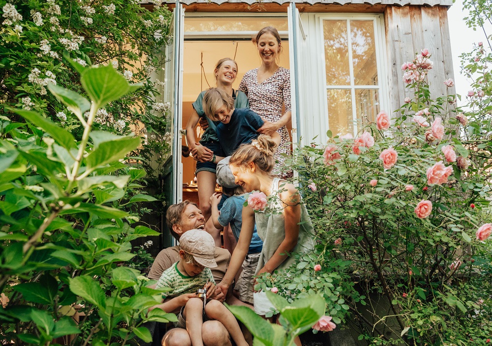 Eine fröhliche sechsköpfige Familie, darunter Eltern und vier Kinder, sitzt an einem sonnigen Tag auf den Stufen eines Holzhauses, umgeben von üppigen Grünpflanzen und blühenden rosa Rosen, und lacht gemeinsam.