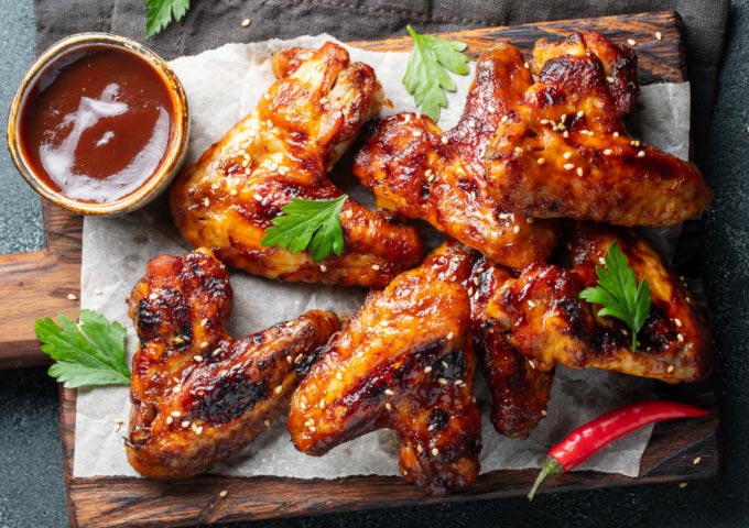 Gegrillte Hähnchenflügel, glasiert mit Barbecue-Sauce, garniert mit Petersilie und Sesam, serviert auf Pergamentpapier mit einer kleinen Schale Sauce und einer roten Chilischote auf einem Holzbrett.