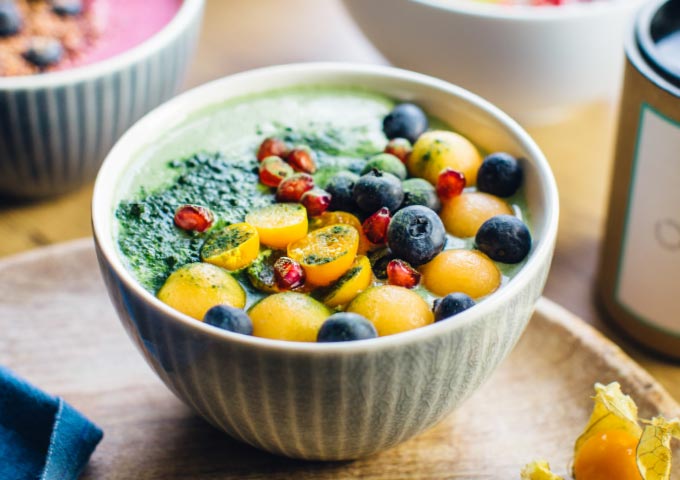 Eine Smoothie-Bowl mit gelben Melonenkugeln, Blaubeeren, in Scheiben geschnittenen goldenen Beeren, Granatapfelkernen und grünem Pulver, serviert in einer grauen Schale auf einer Holzunterlage.