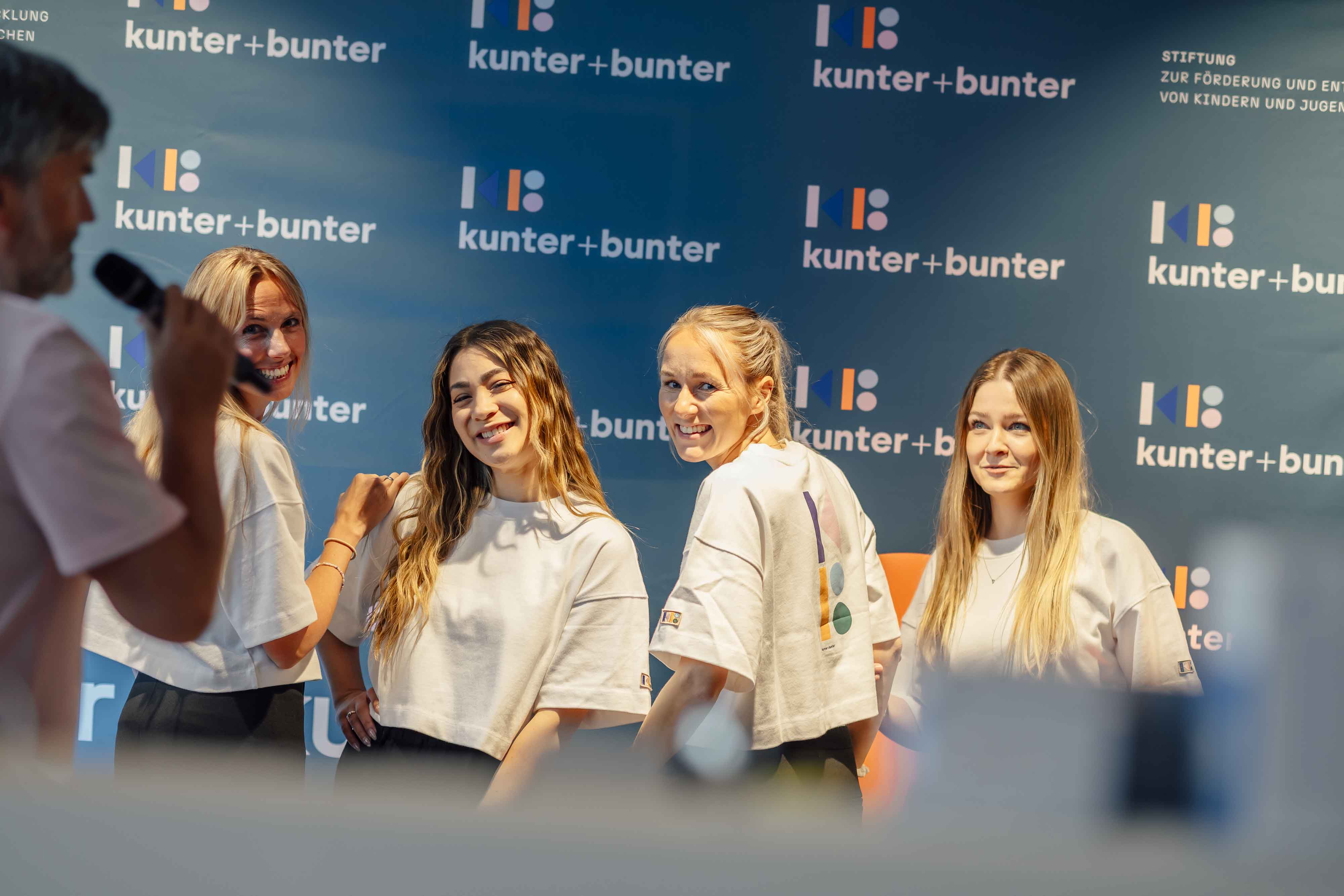 Vier Frauen in weißen Hemden posieren und lächeln in die Kamera vor einem blauen Hintergrund mit kunter+bunter-Logos, während links ein Mann in ein Mikrofon spricht.