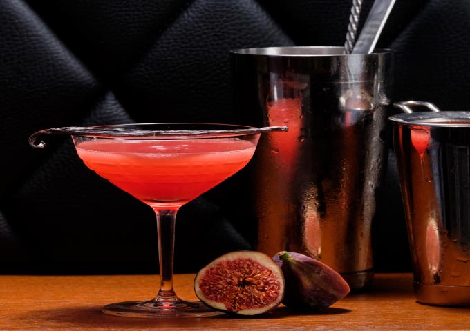Ein rosafarbener Cocktail in einem Coupe-Glas steht auf einer hölzernen Unterlage, garniert mit einem metallenen Rührstab. Frische Feigen, eine ganz und eine aufgeschnitten, liegen vor dem Glas. Im Hintergrund stehen zwei Cocktailshaker aus Metall.