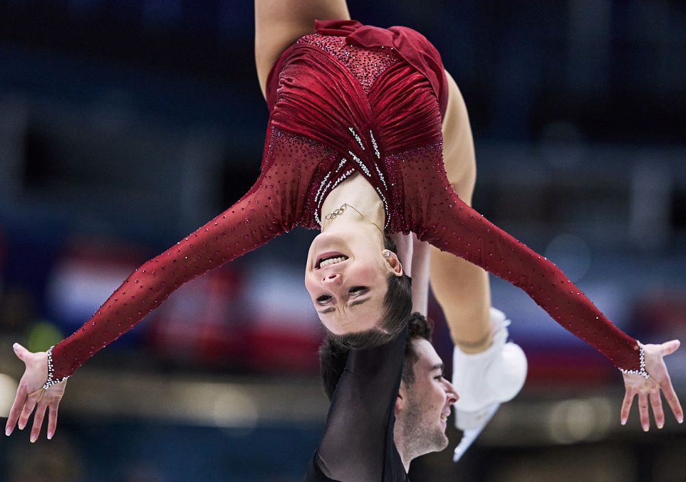 Eine Eiskunstläuferin in einem roten Kostüm wird während einer Hebung mit ausgestreckten Armen kopfüber über den Kopf ihres Partners auf einer Eisbahn gehalten. Der Hintergrund ist unscharf und hebt die dramatische Pose hervor.