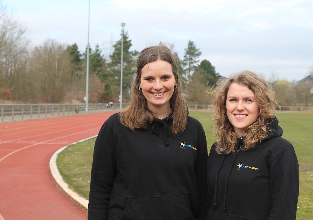 Zwei Frauen in schwarzen Kapuzenpullis stehen lächelnd am Rande einer Laufbahn im Freien, mit Bäumen und einem Feld im Hintergrund unter einem bewölkten Himmel.