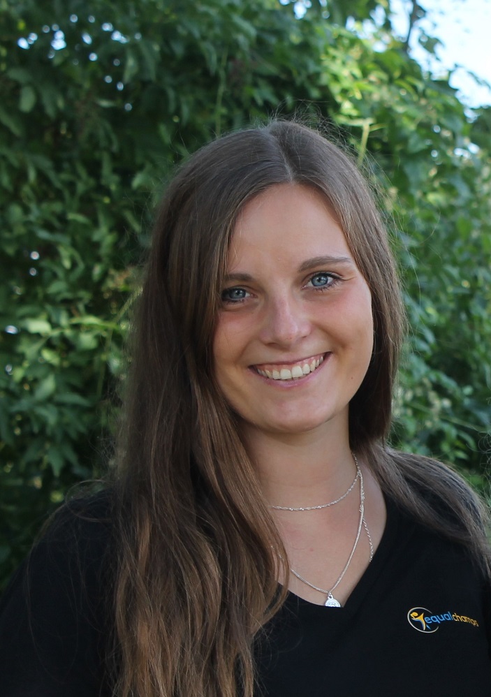 Eine Frau mit langen braunen Haaren und blauen Augen lächelt in die Kamera, während sie im Freien vor grünem Laub steht. Sie trägt ein schwarzes Hemd mit einem gelben Logo und eine silberne Halskette mit einem dreieckigen Anhänger.