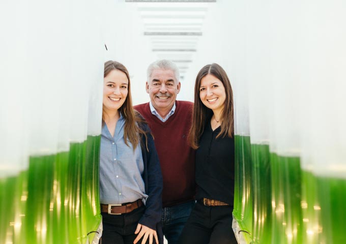 Drei Personen, zwei Frauen und ein älterer Mann, stehen lächelnd zwischen Reihen von grünen Tanks oder Röhren in einer hell erleuchteten, modernen Halle, die möglicherweise mit Wissenschaft oder Landwirtschaft zu tun hat.