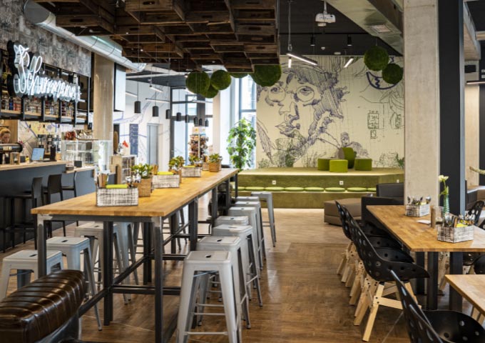 Modernes Café-Interieur mit Holztischen, Metall- und schwarzen Stühlen und einer langen Bar auf der linken Seite. Ein Wandgemälde mit dem Gesicht einer Person und Skizzen ziert die Rückwand. Hängende Pflanzen und warme Beleuchtung schaffen eine gemütliche, trendige Atmosphäre.
