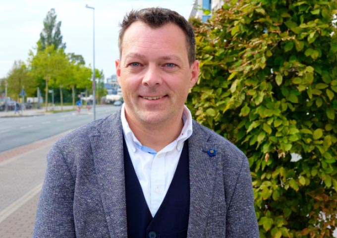 Ein Mann in blauem Blazer und weißem Hemd steht lächelnd auf einem Bürgersteig neben grünen Büschen, mit einer Straße und Bäumen im Hintergrund an einem hellen Tag.