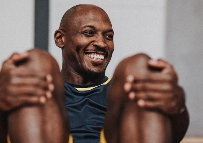 Ein Mann lächelt, während er mit hochgezogenen Knien sitzt, ein dunkles Sporthemd mit gelbem Saum trägt, die Beine hochhält und drinnen entspannt und glücklich wirkt.