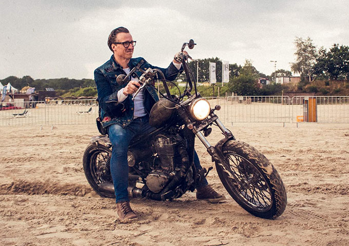 Ein Mann mit Brille, blauer Jacke und Jeans sitzt auf einem Oldtimer-Motorrad auf einer Sandfläche, lächelt und hält eine Kamera in der Hand, im Hintergrund sind Zäune und Bäume zu sehen.