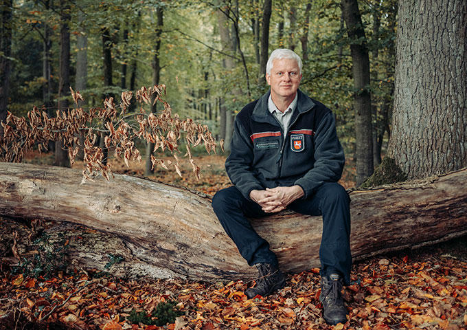 Ein Mann mit grauem Haar, der eine dunkle Jacke und eine schwarze Hose trägt, sitzt auf einem umgestürzten Baumstamm in einem Wald. Herbstblätter bedecken den Boden, und grünes und braunes Laub umgibt ihn.
