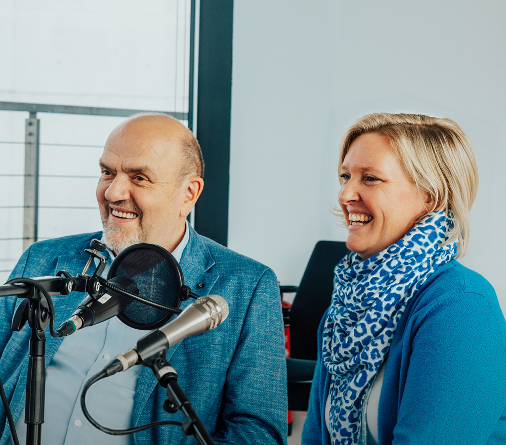 Zwei Personen, ein Mann und Annie Heger, sitzen nebeneinander und lächeln vor Mikrofonen, als würden sie interviewt oder einen Podcast aufnehmen. Beide sind blau gekleidet, und Annie trägt einen blau-weißen Schal.