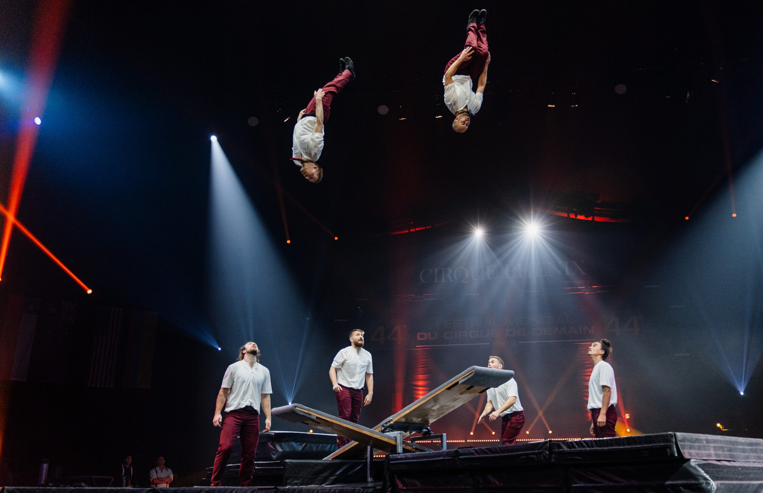 Sechs Akrobaten treten auf der Bühne auf; zwei befinden sich in der Luft und machen Saltos über einer Wippe, während vier von unten zusehen. Die Szene ist mit Bühnenlicht und Scheinwerfern in einem dunklen Theater hell erleuchtet.