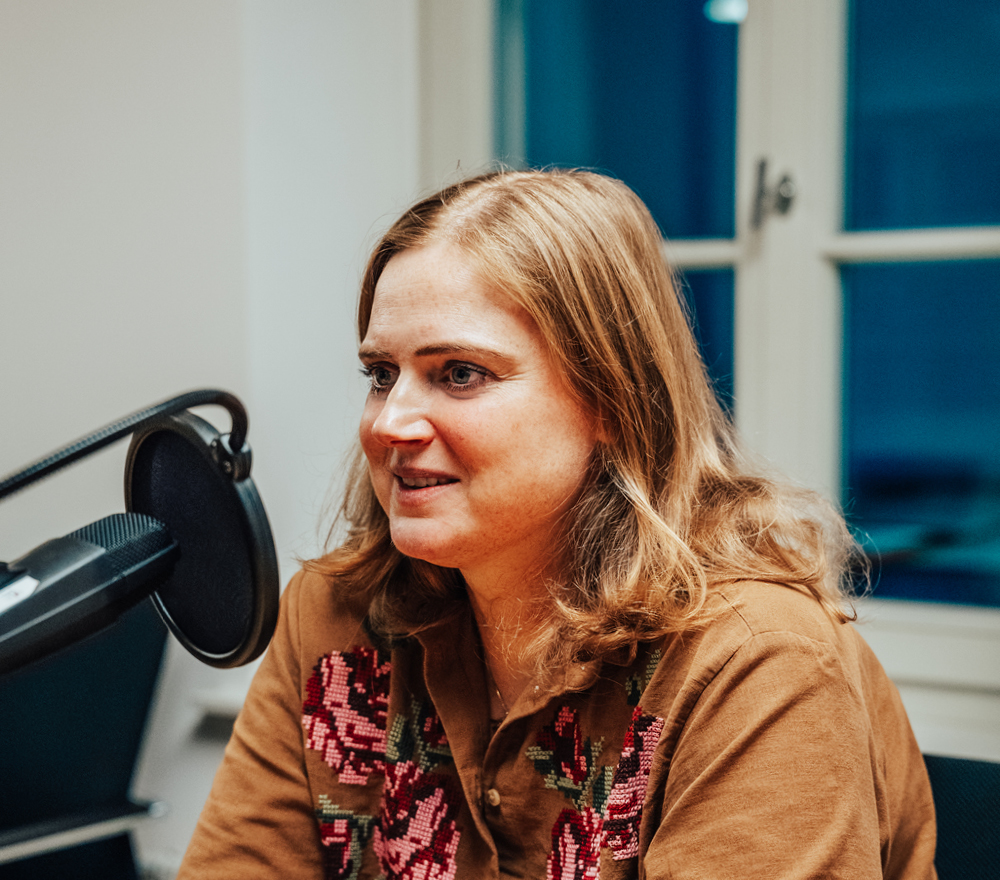 Eine Frau mit schulterlangen hellbraunen Haaren sitzt an einem Tisch vor einem Mikrofon. Sie trägt ein braunes Hemd mit rosa Blumenstickerei und bereitet sich im Nikolausbüro Garrel auf die Weihnachtsfeier vor, mit einem Fenster im Hintergrund.
