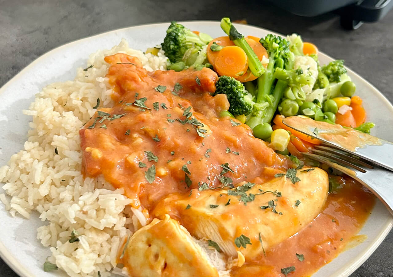 Ein Teller mit weißem Reis, Hähnchenbrust mit roter Soße und Kräutern und einer Beilage aus gemischtem, gedünstetem Gemüse wie Brokkoli, Karotten und Erbsen - perfekt zu Ihren Lieblings-Brotbacken-Rezepten. Gabel und Messer liegen auf dem Teller.