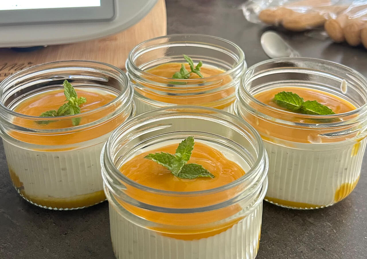 Vier kleine Gläser, gefüllt mit einem cremigen Dessert mit einer leuchtend gelben Soße und frischen Minzblättern, stehen auf einer Arbeitsplatte. Im Hintergrund sind ein Löffel und einige Kekse zu sehen, die perfekt zu den Lieblingsrezepten von Brot Backen passen.