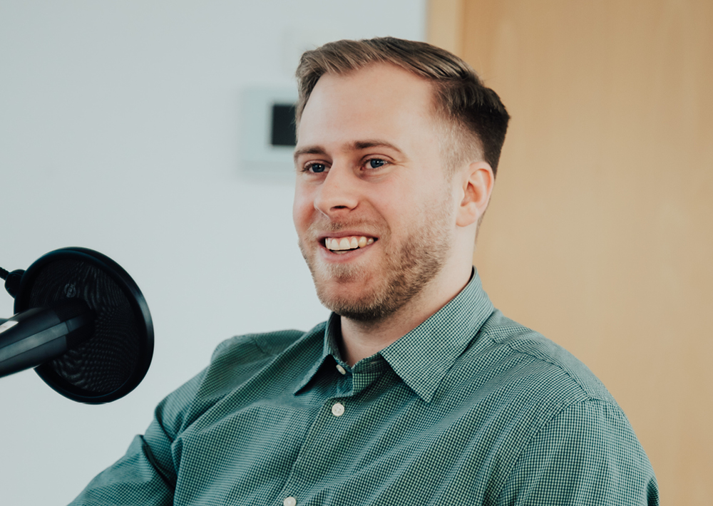 Ein Mann mit kurzen Haaren und gestutztem Bart, der ein grün kariertes Hemd trägt, sitzt da und lächelt, während er in ein schwarzes Mikrofon spricht - vielleicht ein Interview mit Rieke Havertz, Korrespondentin der ZEIT USA. Der Hintergrund ist unscharf, eine Holztür ist zu sehen.