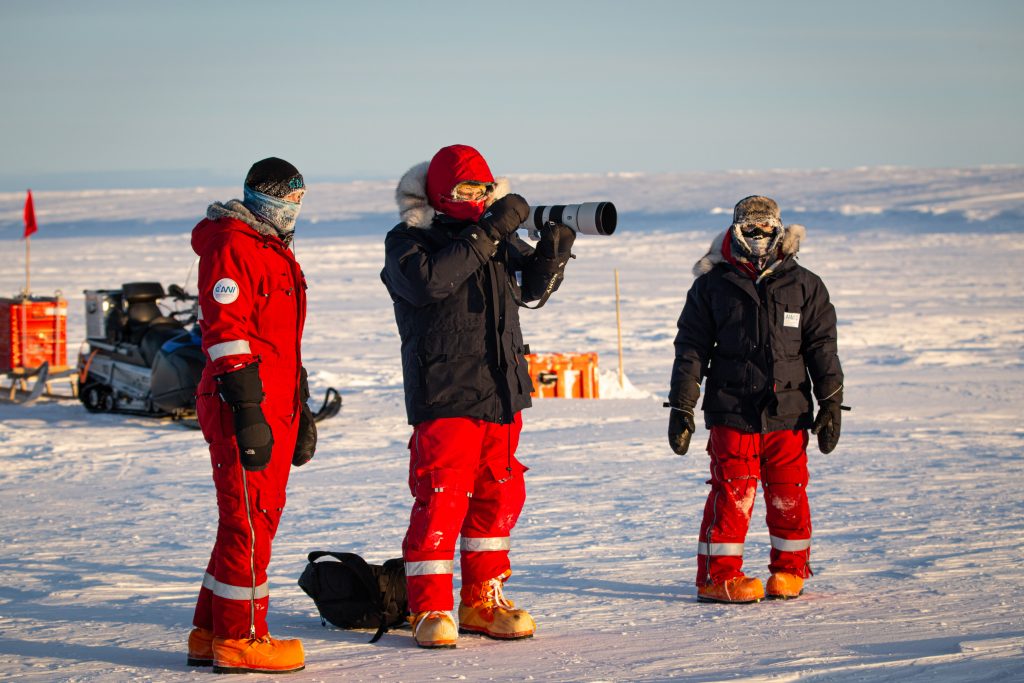 Drei Personen in schwerer Winterausrüstung stehen in einer verschneiten Landschaft; eine von ihnen, möglicherweise Rieke Havertz, Korrespondentin der ZEIT USA, bedient eine Kamera mit einem großen Objektiv, während die anderen zuschauen. Im Hintergrund stehen Motorschlitten und Ausrüstung unter freiem Himmel.