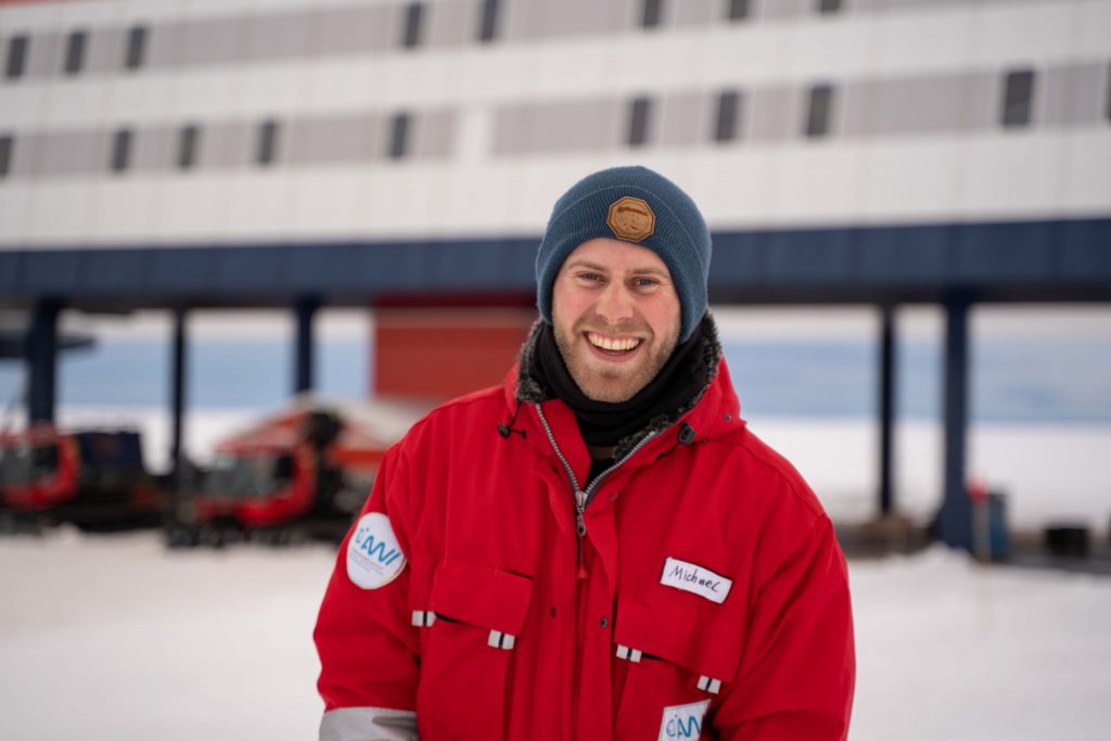 Ein lächelnder Mann in roter Jacke und blauer Mütze steht draußen im Schnee, im Hintergrund ein modernes Gebäude und Fahrzeuge, die an eine Interviewkulisse für Rieke Havertz Korrespondentin ZEIT USA erinnern.